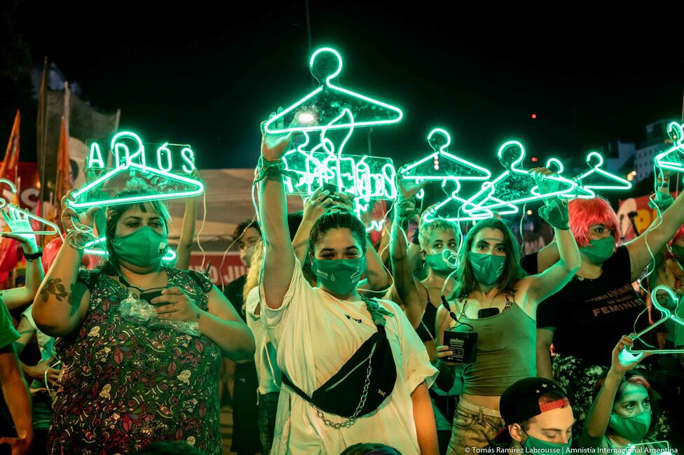 Viele Frauen stehen zusammen, es ist dunkel und sie halten grün leuchtende Kleiderbügel in die Luft. 