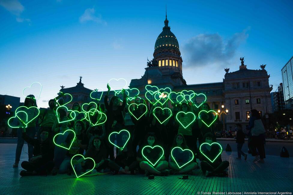 Eine Gruppe von Menschen in der Abenddämmerung halten grün leuchtende Herzen in die Luft. 