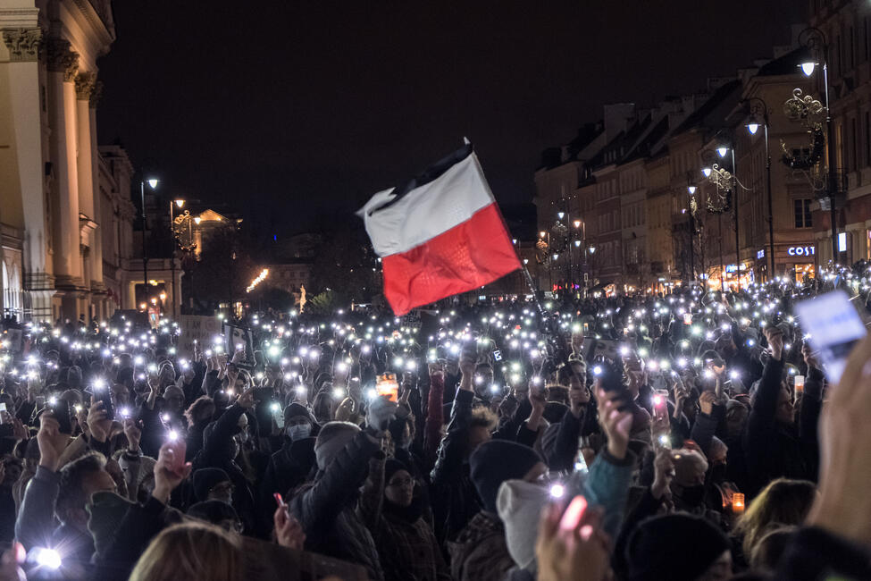 Eine große Menschenmenge, alle halten ihr Handy mit eingeschalteter Taschenlampe hoch. In der Mitte wird eine polnische Nationalflagge hochgehalten. 