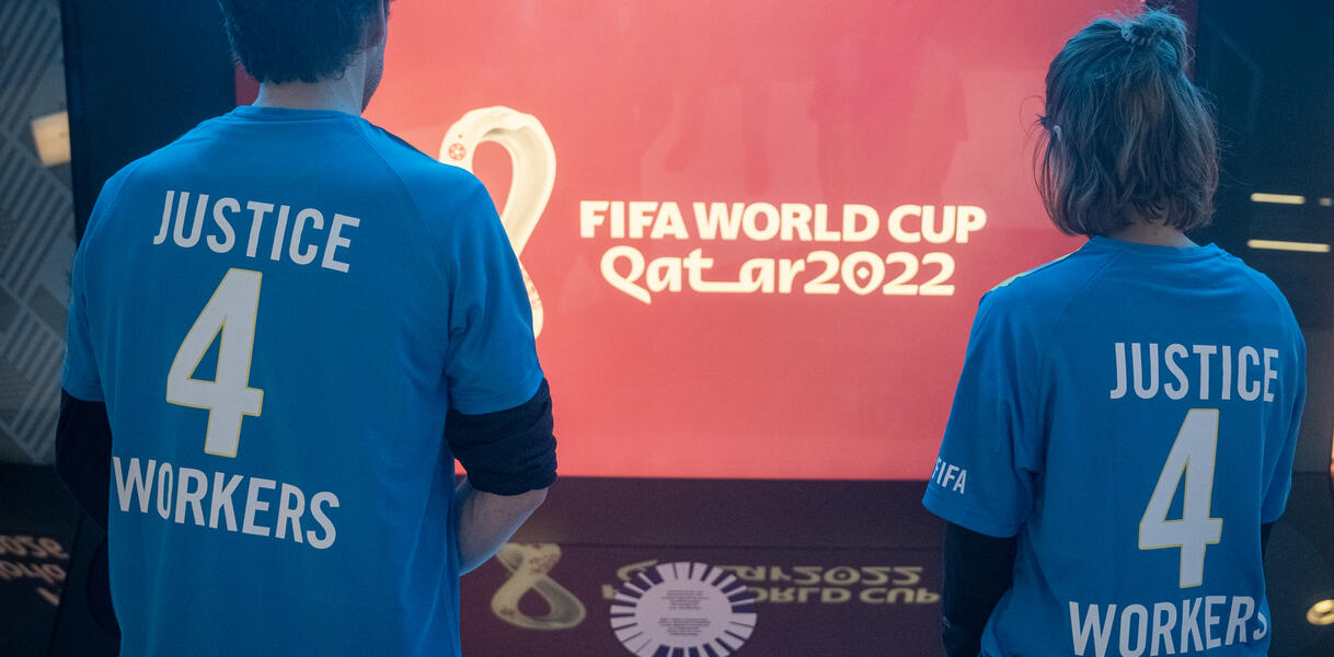Das Bild zeigt zwei Personen von hinten, die auf einen Bildschirm schauen. Darauf steht "FIFA World Cup"