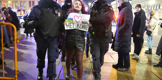 Das Bild zeigt, wie mehrere Polizisten eine junge Frau abführen. Sie trägt ein Schild mit der Aufschrift "Kein Krieg". 