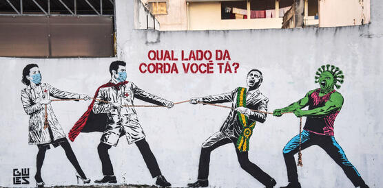 Ein Grafitti im brasilianischen Sao Paolo, das Corona thematisiert: Menschen stehen an einem Seil, Tauziehen, auf der einen Seite medizinisches Personal mit Mundnasenschutzmasken, auf der anderen Seite des Seils der ehemalige Präsident Jair Bolsonaro und das Corona-Virus in Menschengestalt.