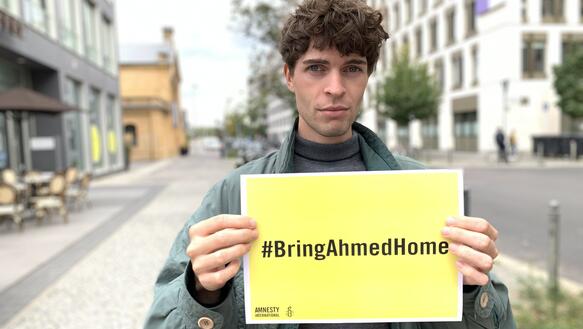 Ein junger Mann steht auf der Strasse und hält ein gelbes Schild in die Kamera. Darauf steht der Aufruf: "#BringAhmedHome"