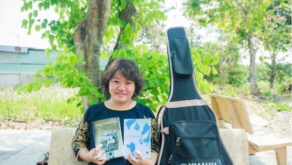 Eine lächelnde Frau mit kinnlangen dunklen Haaren sitzt auf einer Parkbank und zeigt zwei vietnamesische Bücher in die Kamera. Neben ihr ist eine Gitarrentasche.