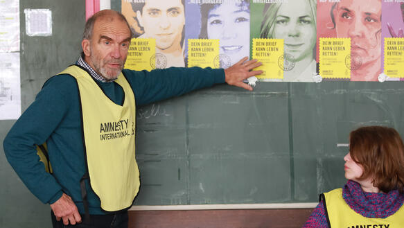 Ein älterer Mann mit Amnesty-Weste steht in einem Klassenzimmer vor einer Tafel und zeigt mit seiner linken Hand auf dort angebrachte Amnesty-Poster. Ein Kind in der rechten Ecke des Bildes schaut ihn an.