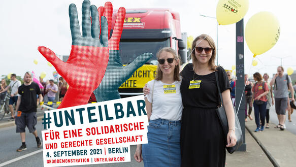 Zwei Frauen lächeln in die Kamera und halten gelbe Luftballons. Sie stehen vor einem großen Truck mit gelben Banner. Auf dem Foto ist ein Logo mit zwei Händen platziert sowie der Text: #Unteilbar I Für eine solidarische und gerechte Gesellschaft | 4. September 2021 | Berlin | Großdemonstration | unteilbar.org