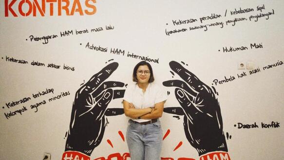 Eine junge Frau steht vor einer bemalten Wand. Sie verschränkt ihre Arme und lächelt. Auf der Wand sind zwei Hände in Handschellen gemalt, sowie einige Schriftzüge, darunter auch der Name der Organisation KontraS.