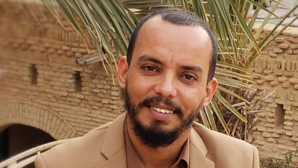 Portraitfoto eines lächelnden Mannes, der einen beigen Anzug und ein braunes Hemd trägt, im Hintergrund sind Palmblätter