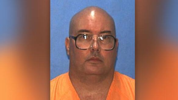 Porträtfoto von Donald Dillbeck, dessen Kopf kahlrasiert ist und der eine Brille und Gefängniskleidung trägt.