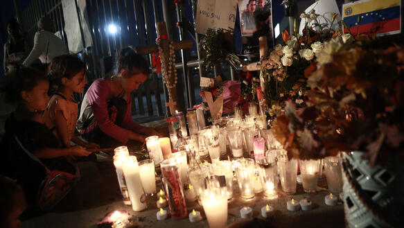 Das Bild zeigt Kinder die in der Dunkelheit vor vielen brennenden Kerzen auf dem Boden knien. Hinter den Kerzen stehen mehrere Blumensträße, Kreuze und ein Bild, das einen Mann mit einem Kind zeigt.