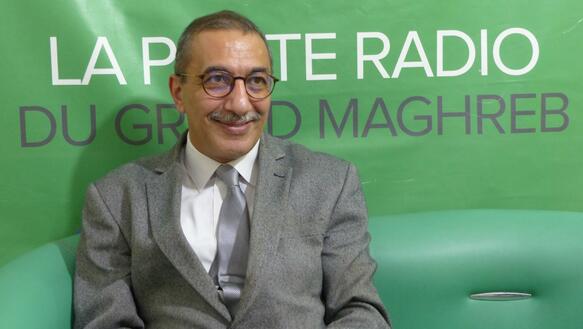 Das Foto zeigt Ihsane El Kadi lächelnd im Anzug mit Krawatte vor einem Banner sitzen, auf dem unter anderem die Wörter "Radio" und "Maghreb" stehen.