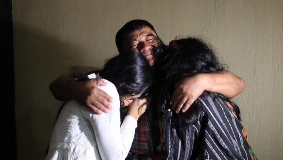 Das Bild zeigt einen Mann, der die Augen geschlossen hat, grinst und zwei Frauen umarmt.