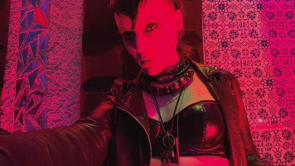 Eine junge Frau mit Punkfrisur, Lederoutfit, Ketten um den Hals im Ambiente eines Clubs.