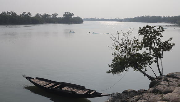Ein sehr breiter Fluss, inmitten dessen Fischernetze aufgespannt sind; am Ufer dümpelt ein Holzboot, das an einem kleinen Baum am Ufer mit einem Seil angebunden ist.