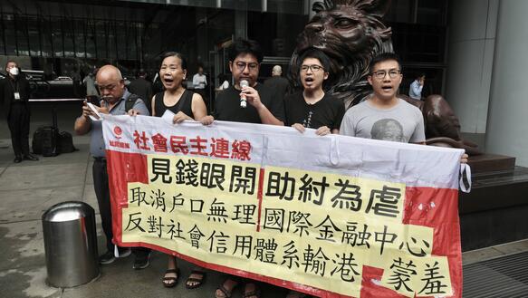 Fünf Personen halten ein Banner hoch mit chinesischer Schrift und stehen vor dem Eingang zu einem Gebäude.