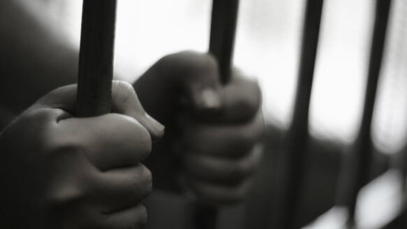 Zwei Hände umfassen die Gitterstäbe einer Gefängniszelle.