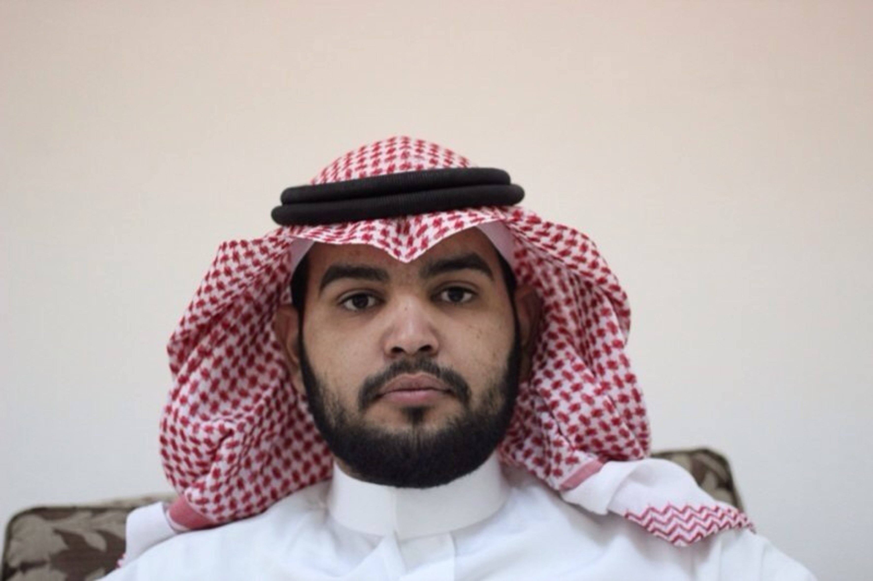 Porträtfoto von Abdulaziz al-Shubaily, der ernst in die Kamera blickt. Er trägt eine traditionelle Kopfbedeckung: Ein kariertes Kopftuch.