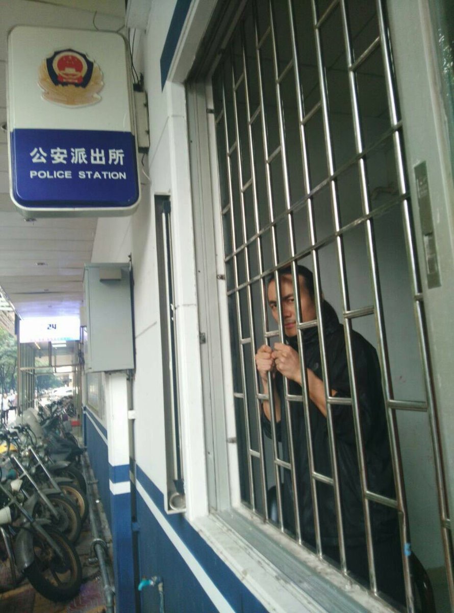 Zhen Jianghua ist hinter Gittern, seine Hände an den Gitterstäben. An der Außenwand ein Schild: Polizeiwache.