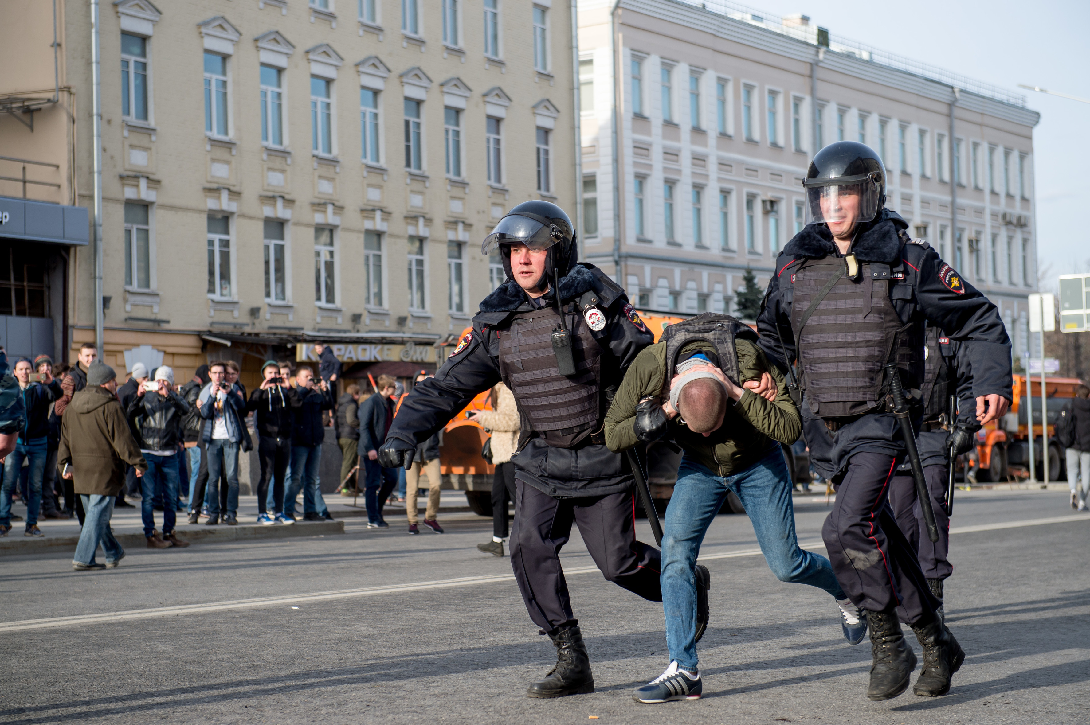 Zwei Polizisten mit Schutzwesten und Helmen auf den Köpfen zerren einen Mann zwischen sich mit, im Hintergrund sieht man eine Menschenmenge, einige von ihnen fotografieren die drei Männer