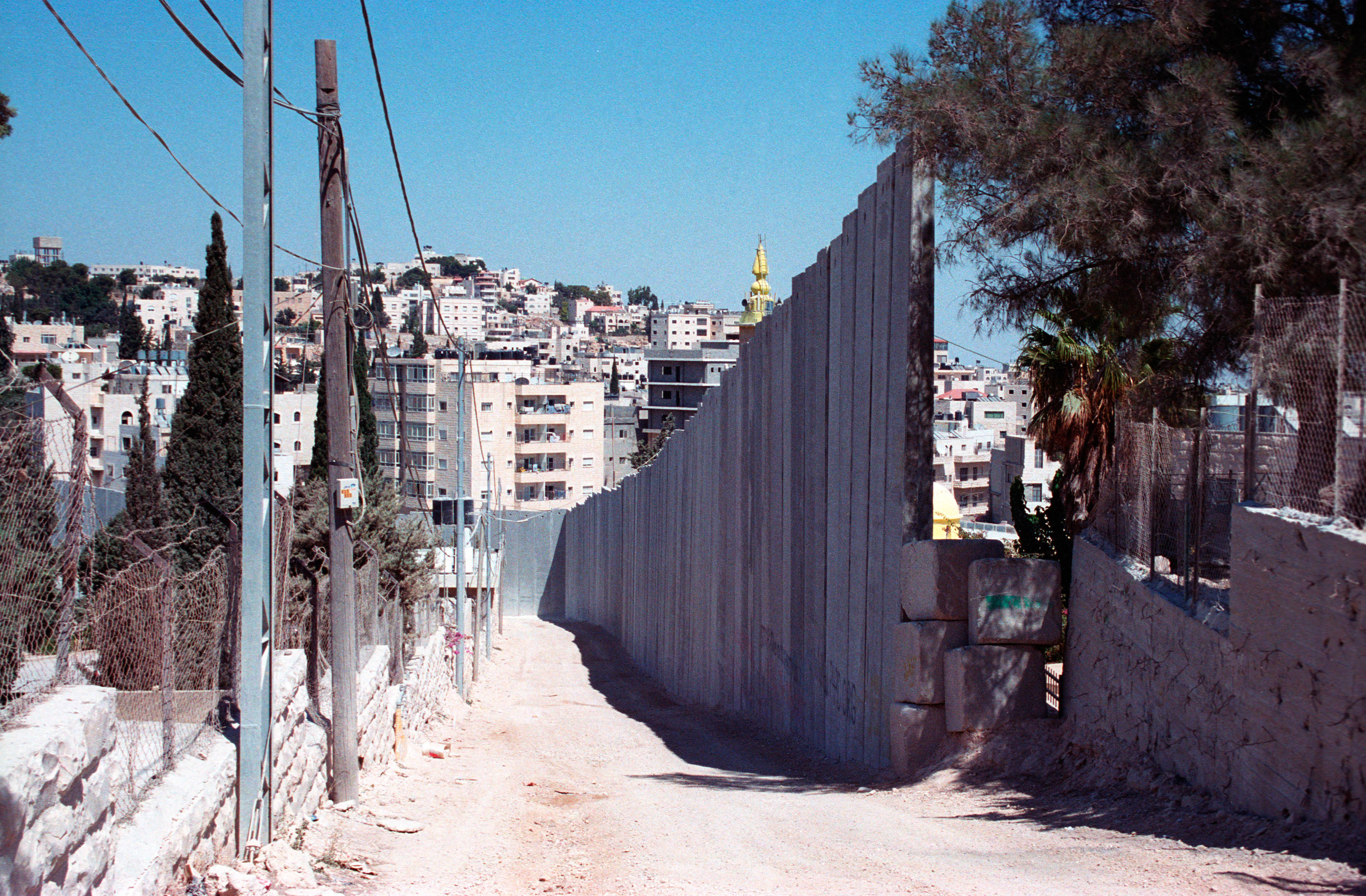 Eine ungepflasterte Straße entlang einer Mauer, im Hintergrund Häuser