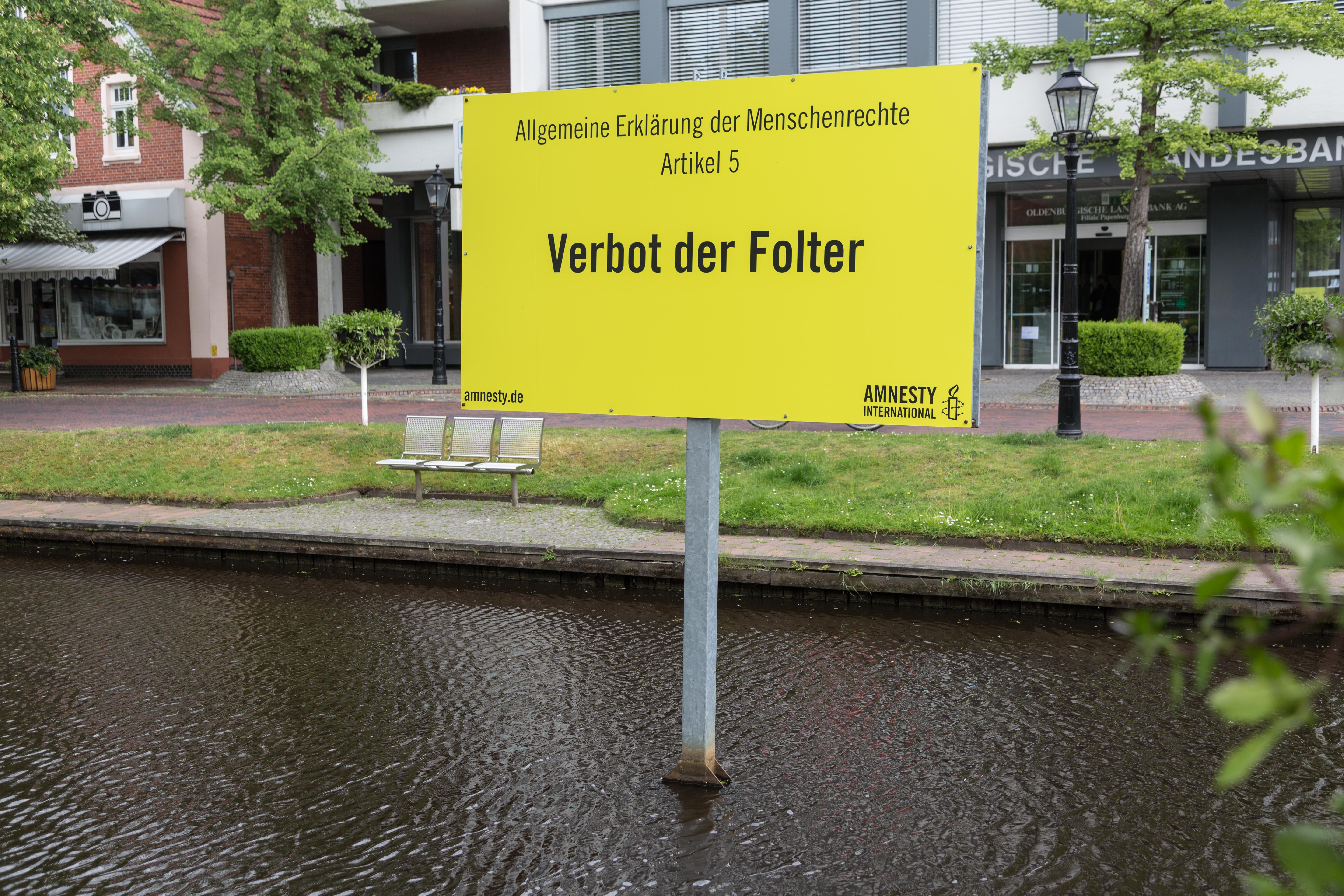 Ein großes gelbes Schild ist aufgestellt in einem innerstädtischen Wasserkanal. Auf ihm steht der Artikel aus der "Allgemeinen Erklärung der Menschenrechte: "Verbot der Folter" 