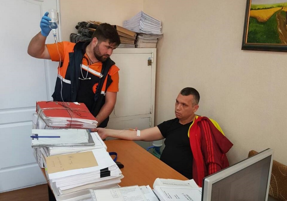 Ein Mann mit einer Kanüle im Arm sitzt, ein anderer mit einem Tropf in der Hand steht daneben