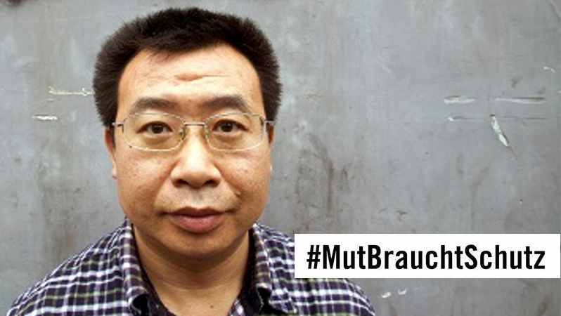 Ein Mann chinesischer Herkunft mit Brille steht vor einer grauen Wand und schaut ernst in die Kamera
