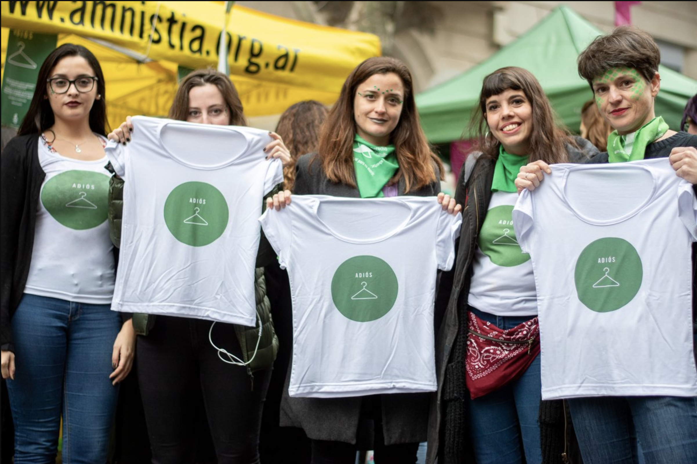Fünf junge Frauen halten weiße T-Shirts hoch auf denen ein grüner Kreis mit einem weißen Kleiderhaken zu sehen ist 