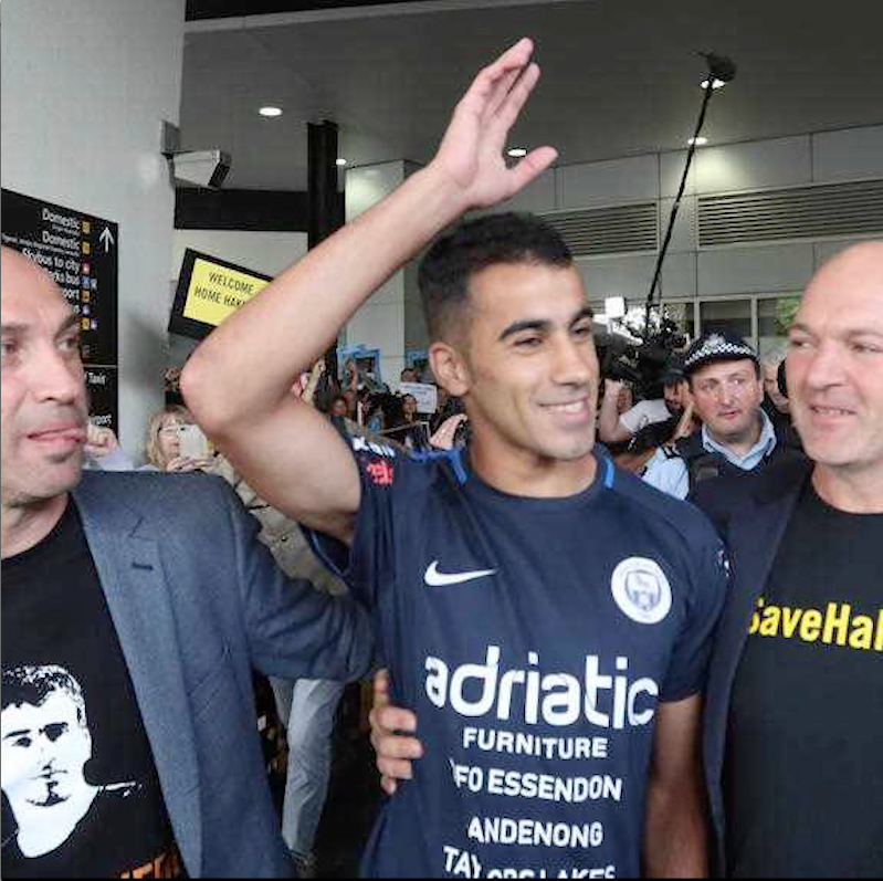 Hakeem Ali al-Araibi steht lachend in der Mitte einer Personengruppe und hält den Arm hoch. Im Hintergrund sind Fluchhafenleitsysteme zu erkennen.