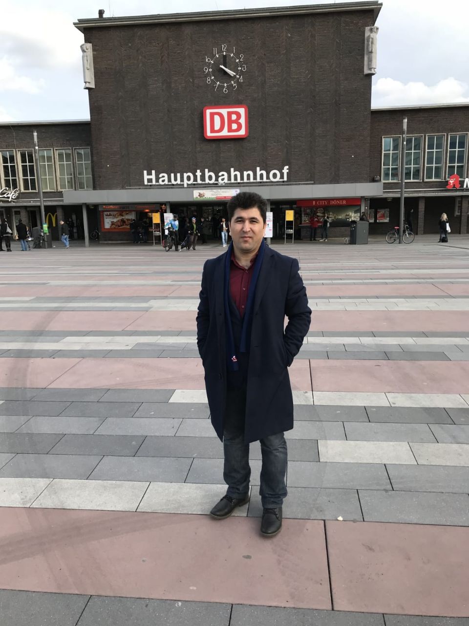 Ein Mann mit Jeans und schwarzem Mantel steht auf dem Vorplatz eines Bahnhofs. Er schaut in die Kamera. Im Hintergrund ist das Bahnhofsgebäude zu sehen.