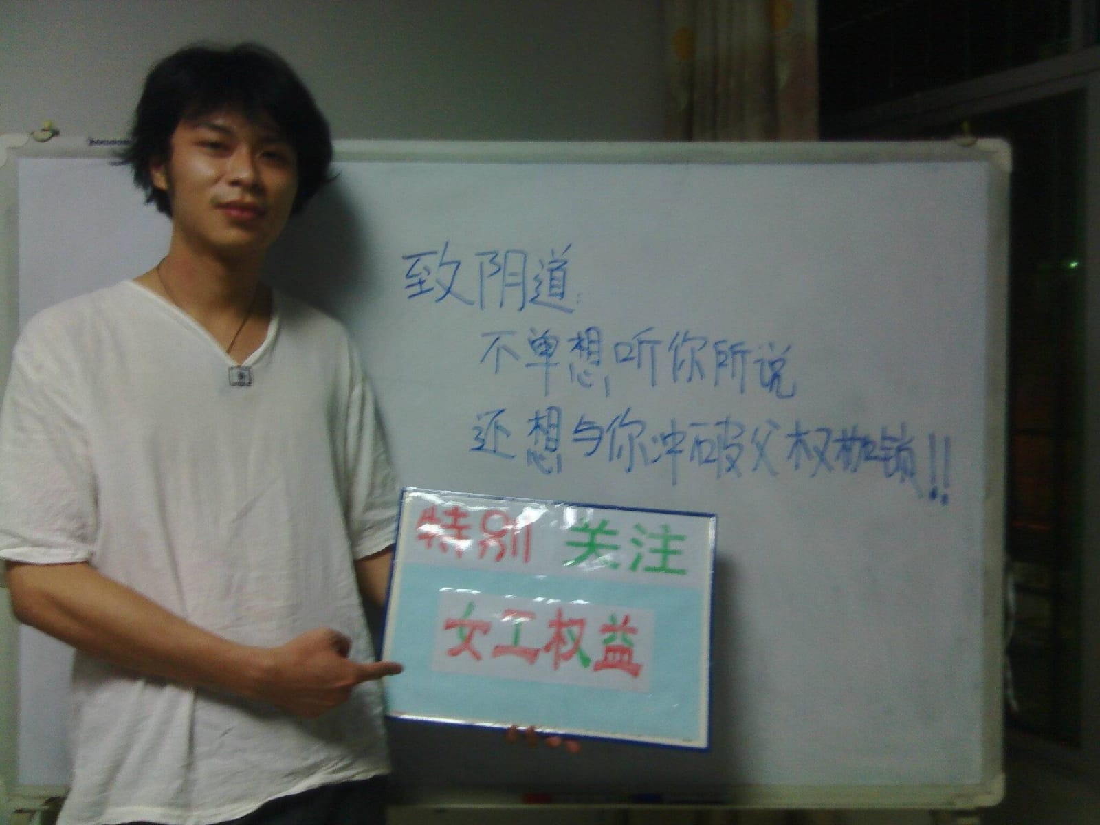 Ein junger Mann aus China steht in einem weissen T-Shirt vor einer Tafel mit chinesischen Zeichen und hält eine Botschaft auf Chinesisch in die Kamera