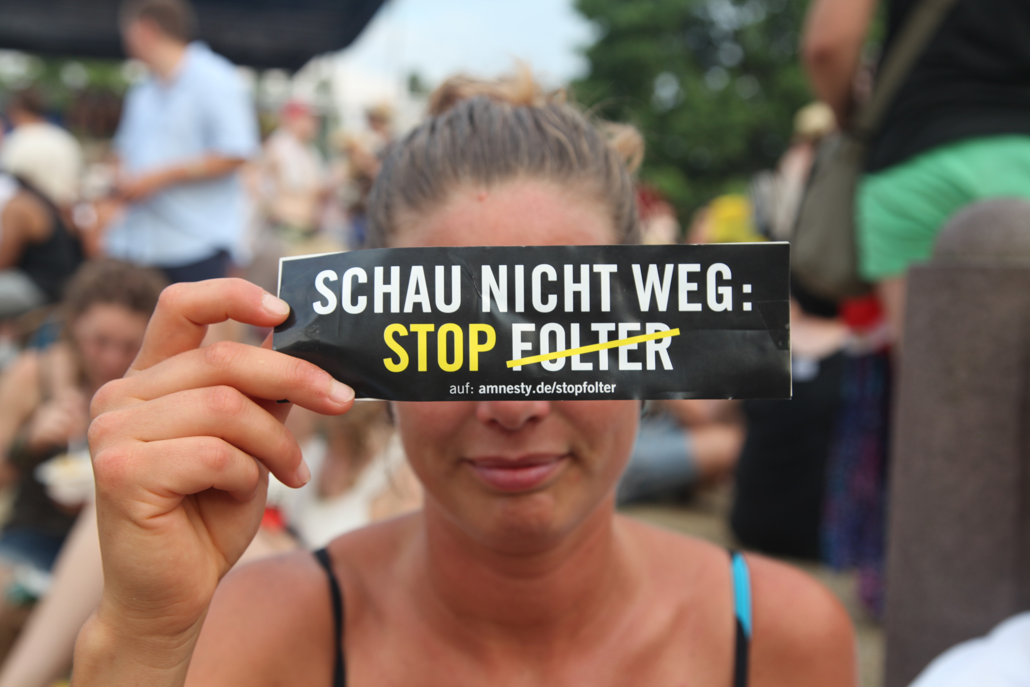 Eine junge Frau hält sich ein schmales Schild vor die Augen auf dem zu drauf steht: "Schau nicht weg. Stop Folter". Folter ist dabei durchgestrichen.