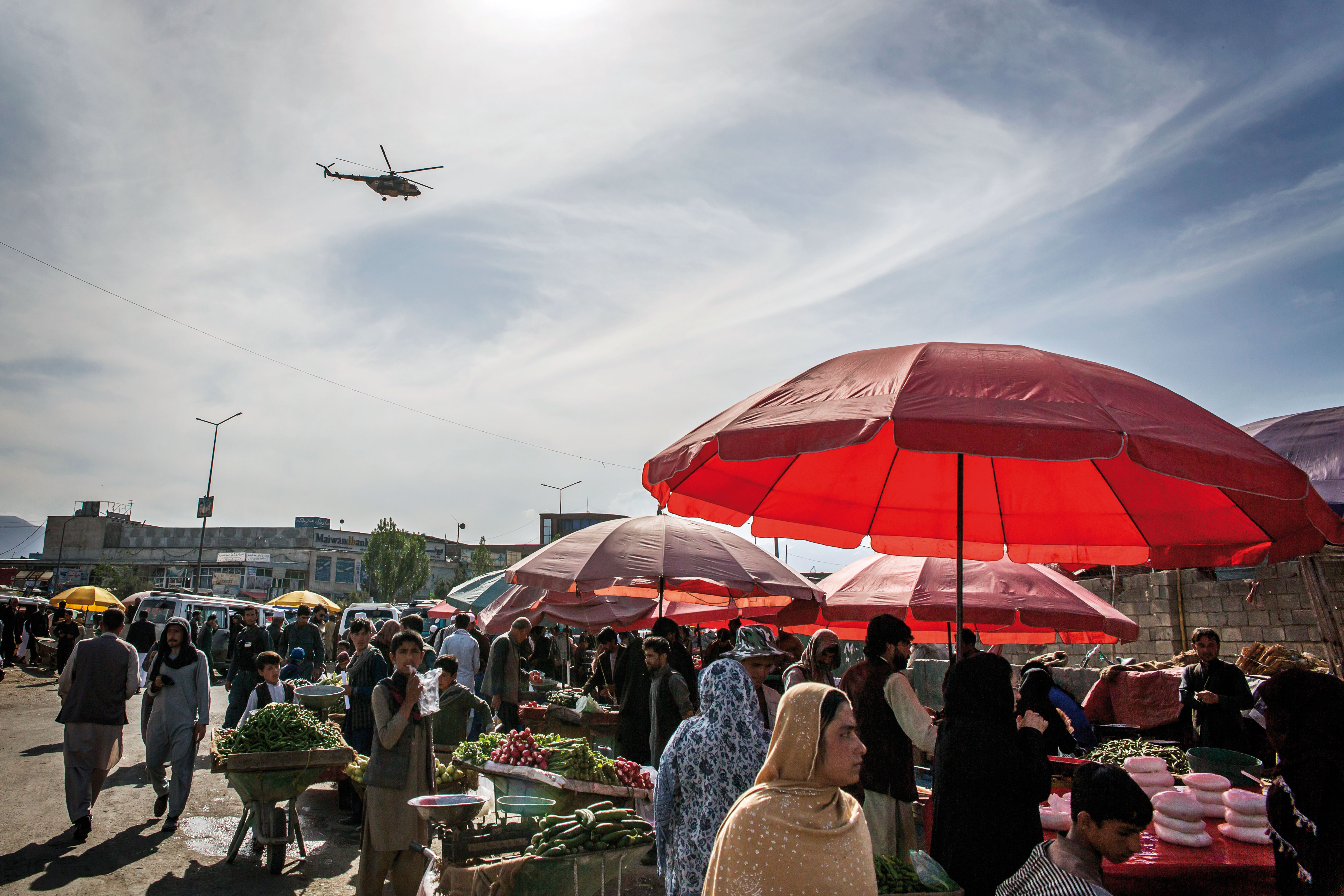 Ein Marktplatz mit Menschen, im Vordergrund ein Sonnenschirm, am Himmel ein Hubschrauber