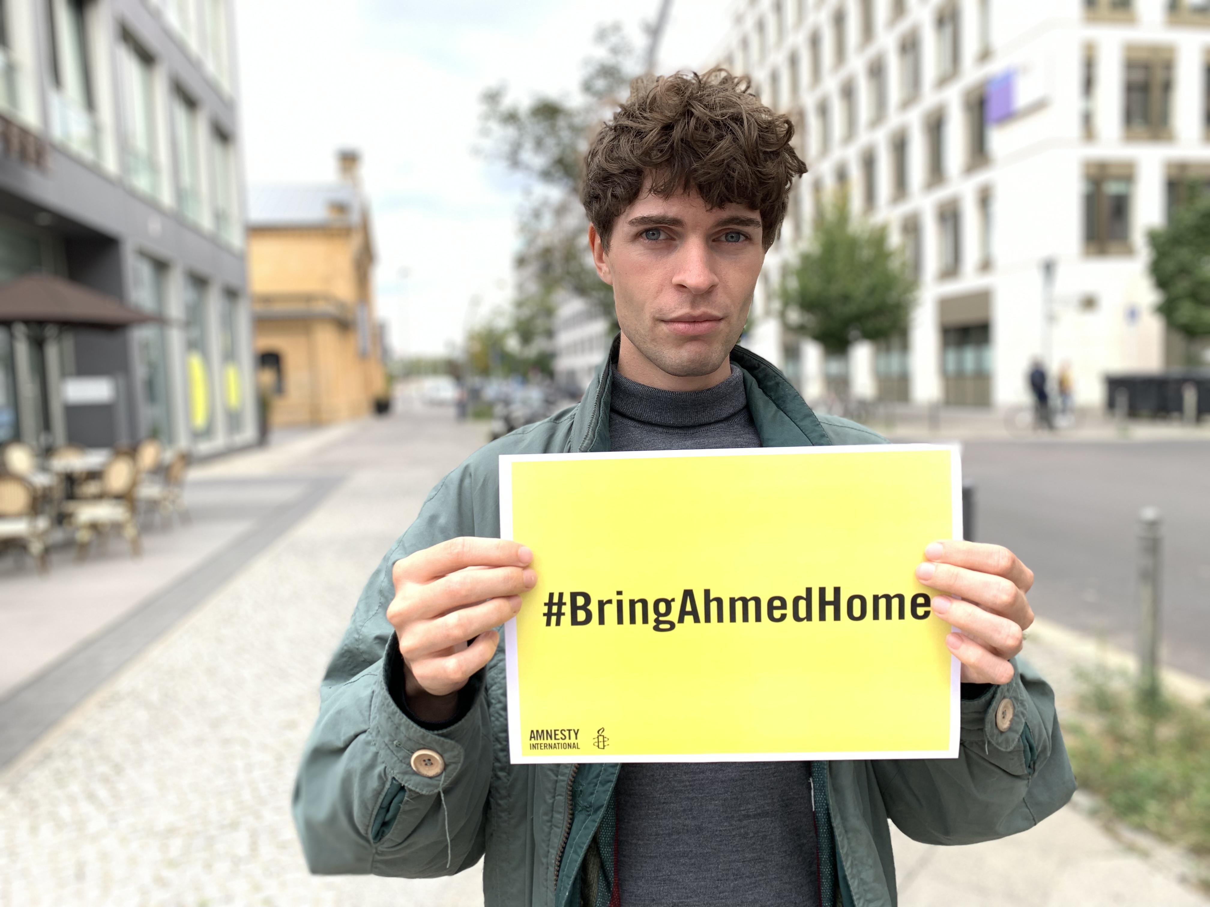 Ein junger Mann steht auf der Strasse und hält ein gelbes Schild in die Kamera. Darauf steht der Aufruf: "#BringAhmedHome"