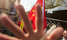 Eine Hand verdeckt teilweise die Kamera. Im Hintergrund befinden sich protestierende Menschen, welche die chinesische Flagge schwenken. 