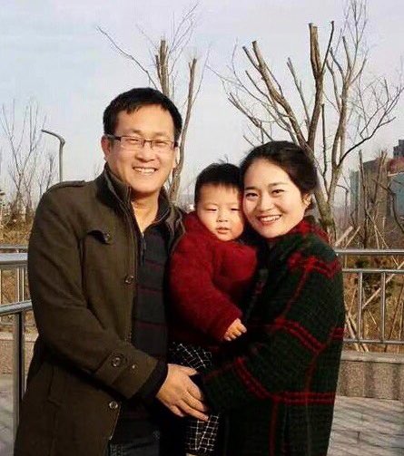 Ein junger chinesischer Mann mit Brille und eine junge chinesische Frau mit naxch hinten gekämmten Haar halten gemeinsam ein kleines Kind auf dem Arm und lächeln in die Kamera.