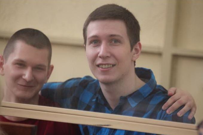 Zwei junge Männer mit kurz geschorenen Haaren umarmen sich und lächeln in die Kamera. Yan Sidorov (links) trägt einen roten Pulli und Vladislav Mordasov (rechts) ein blau-schwarz kariertes Hemd.