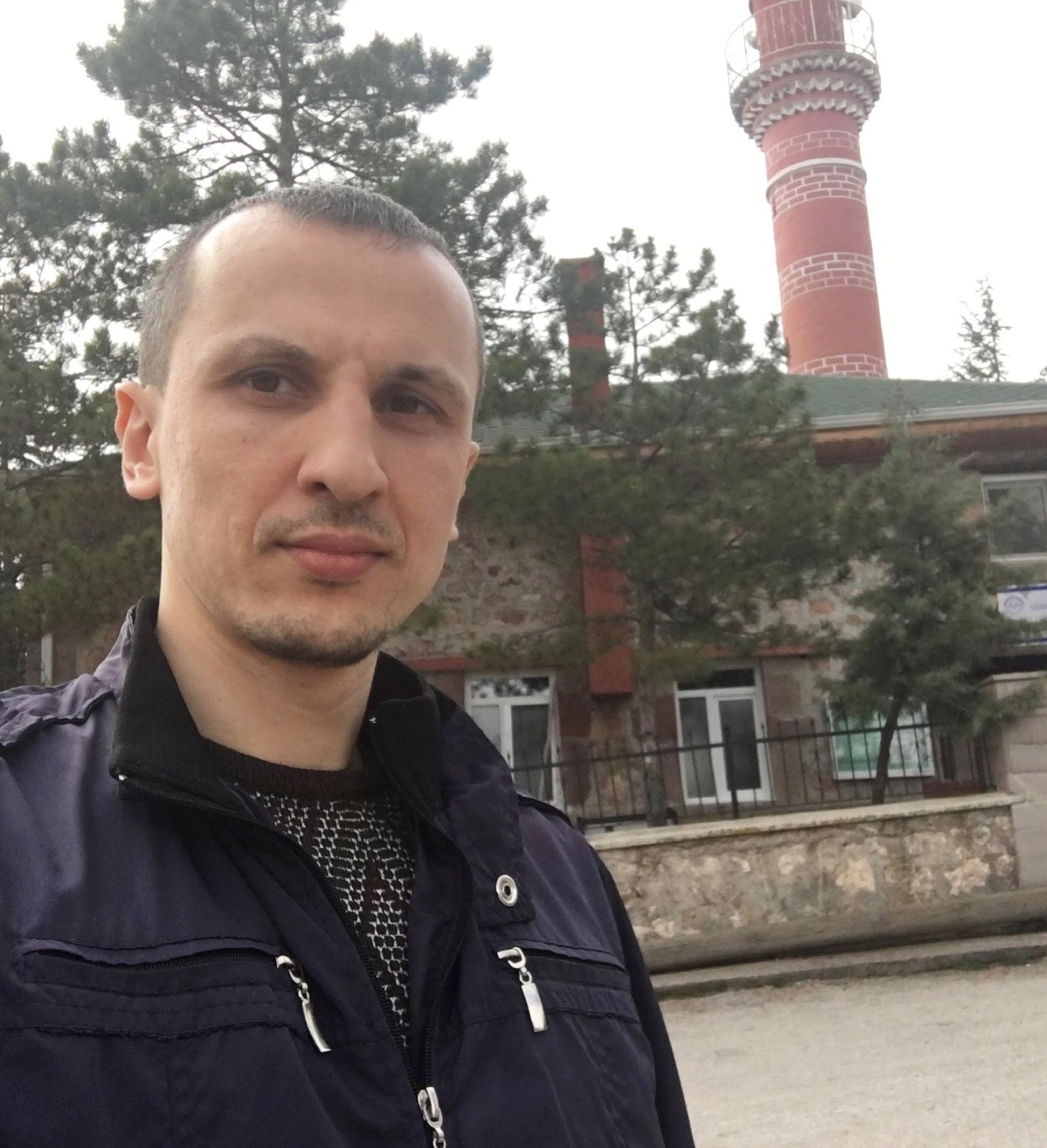 Junger Mann mit ernstem Blick in blauer Jacke. Im Hintergrund ist ein Gebäude zu sehen.