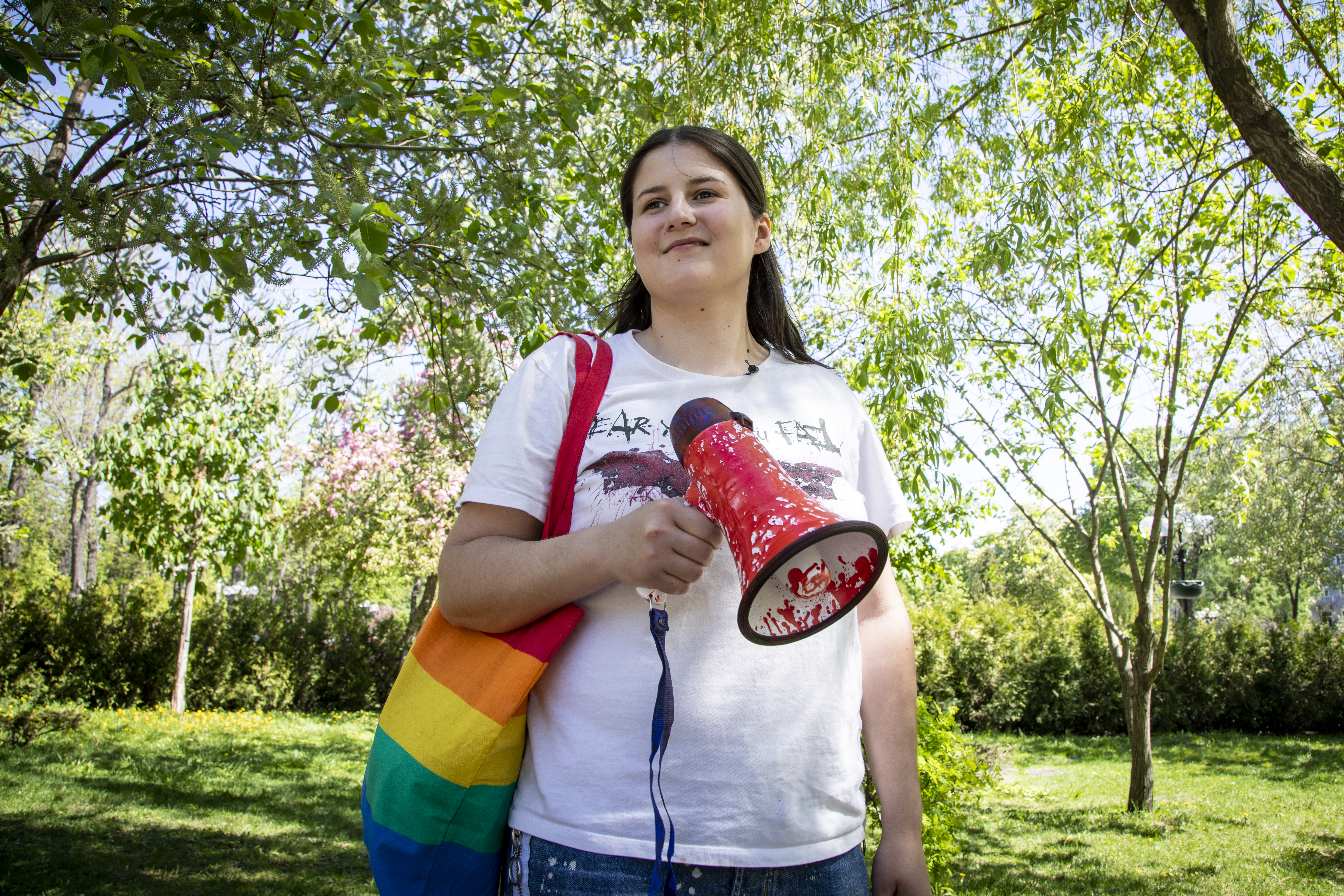 Frau mit Megaphon und Tasche in Regenbogenfarben steht an einem sonnigen Tag in einem Park