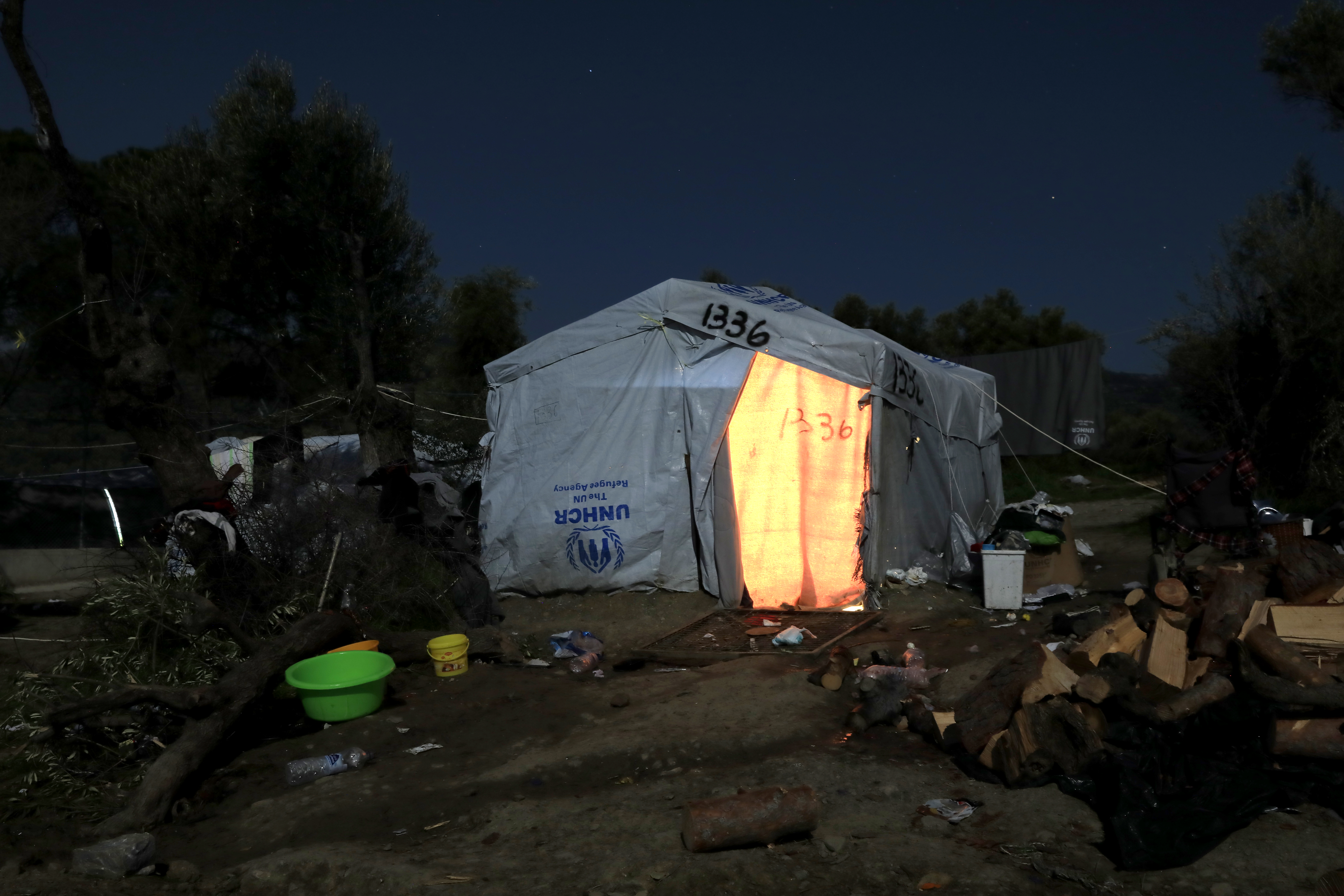In der Mitte des Bildes ist ein großes weißes UNHCR-Zelt zu sehen, aus dem es hell leuchtet, rings herum liegt Müll und verschiedene Gegenstände. Im Hintergrund sind Bäume zu sehen, es ist Nacht und dunkel.