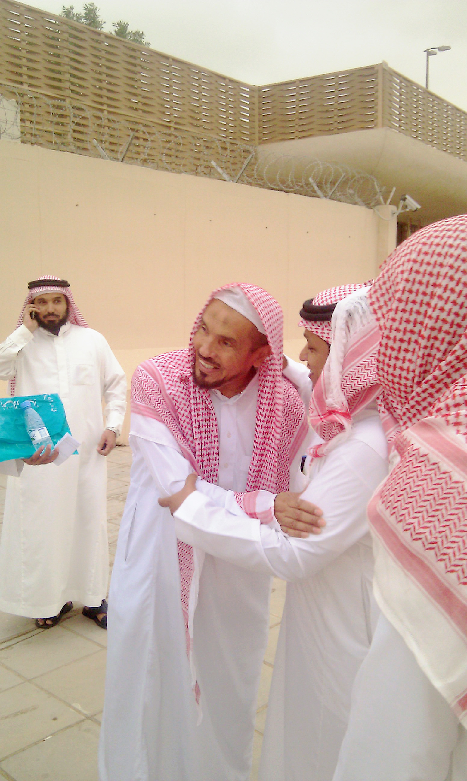 Vier Männer stehen vor einem einstufigem Gebäude. Sie tragen alle weiße saudi-arabischen Gewändern und rot-weißen Kopfbedeckungen. 