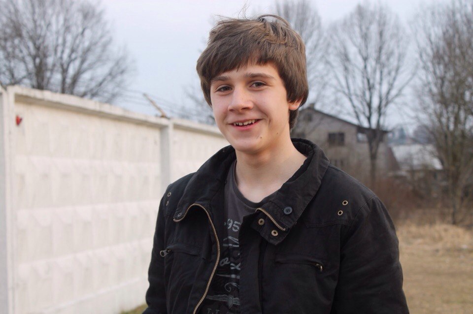 Ein Jugendlicher mit dunklem Haar steht vor einer weißen Mauer und lächelt in die Kamera