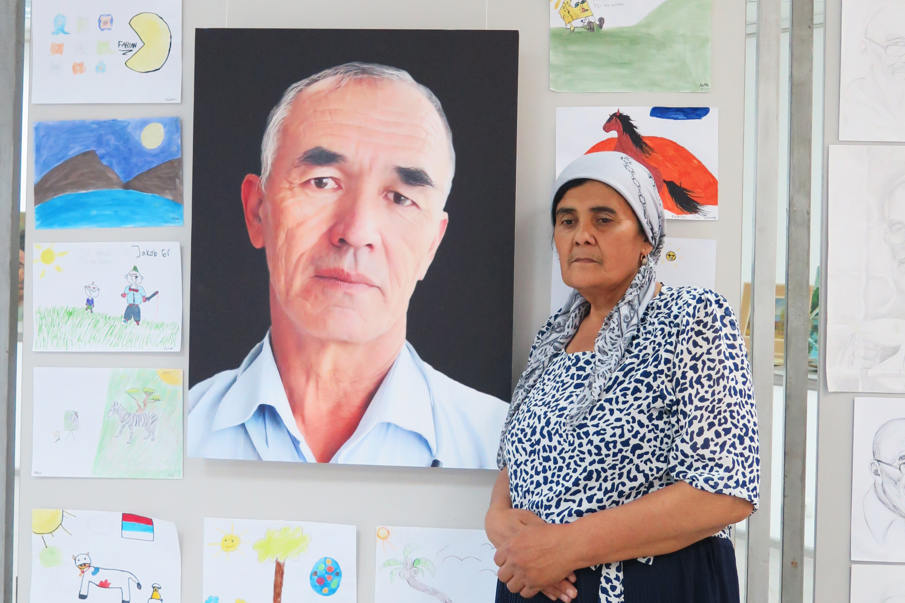 Gezeichnetes Porträtbild von Azimjan Askarov, das an einer Wand hängt. Um sein Porträt hängen Kinderzeichnungen mit verschiedenen Motiven, wie Pferde, Sonne und Mond. Daneben steht eine Frau, die traurig nach unten blickt.