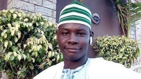 Porträtfoto von Yahaya Sheriff-Aminu, aufgenommen vor einer Hecke vor einen Gebäude. Er trägt eine traditionelle Kopfbedeckung und ein traditionelles Gewand und lächelt in die Kamera.