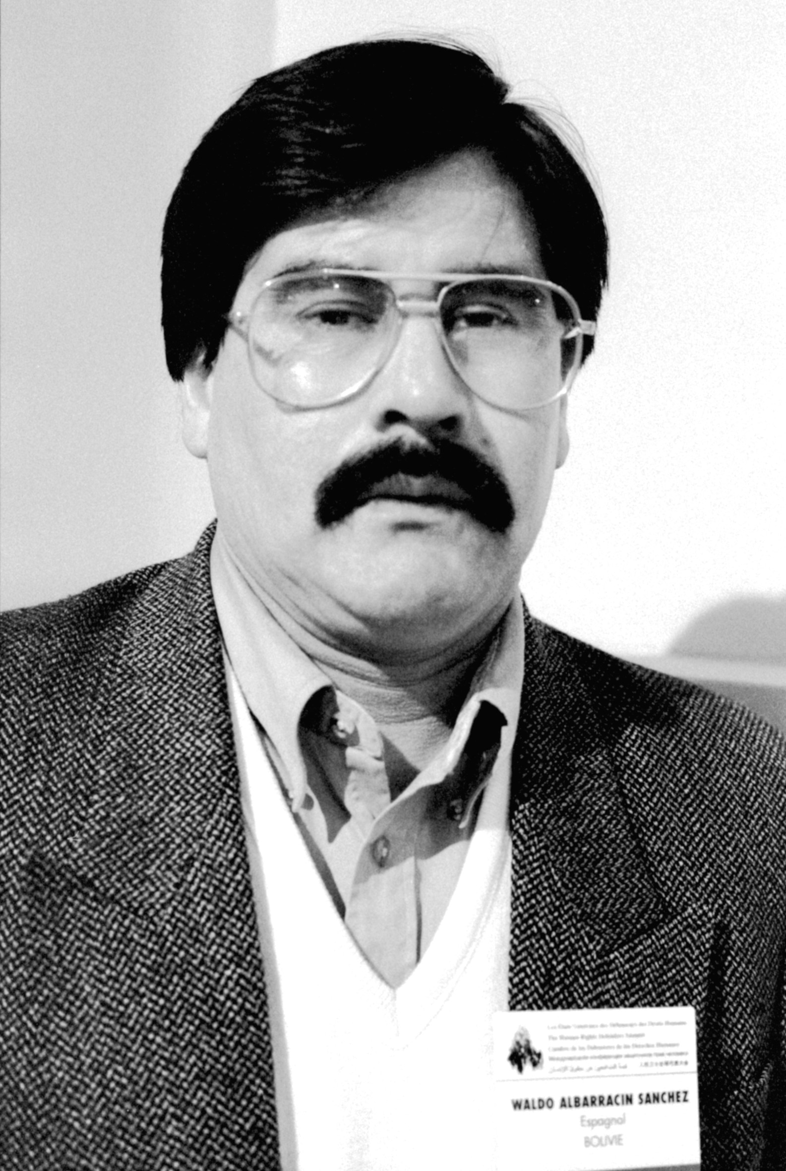 Ein schwarz-weiss Foto von einem Mann mit dunklen Haaren und Schnurrbart. Er trägt eine grosse Pilotenbrille und ein weisses Hemd mit dunklem Jackett.