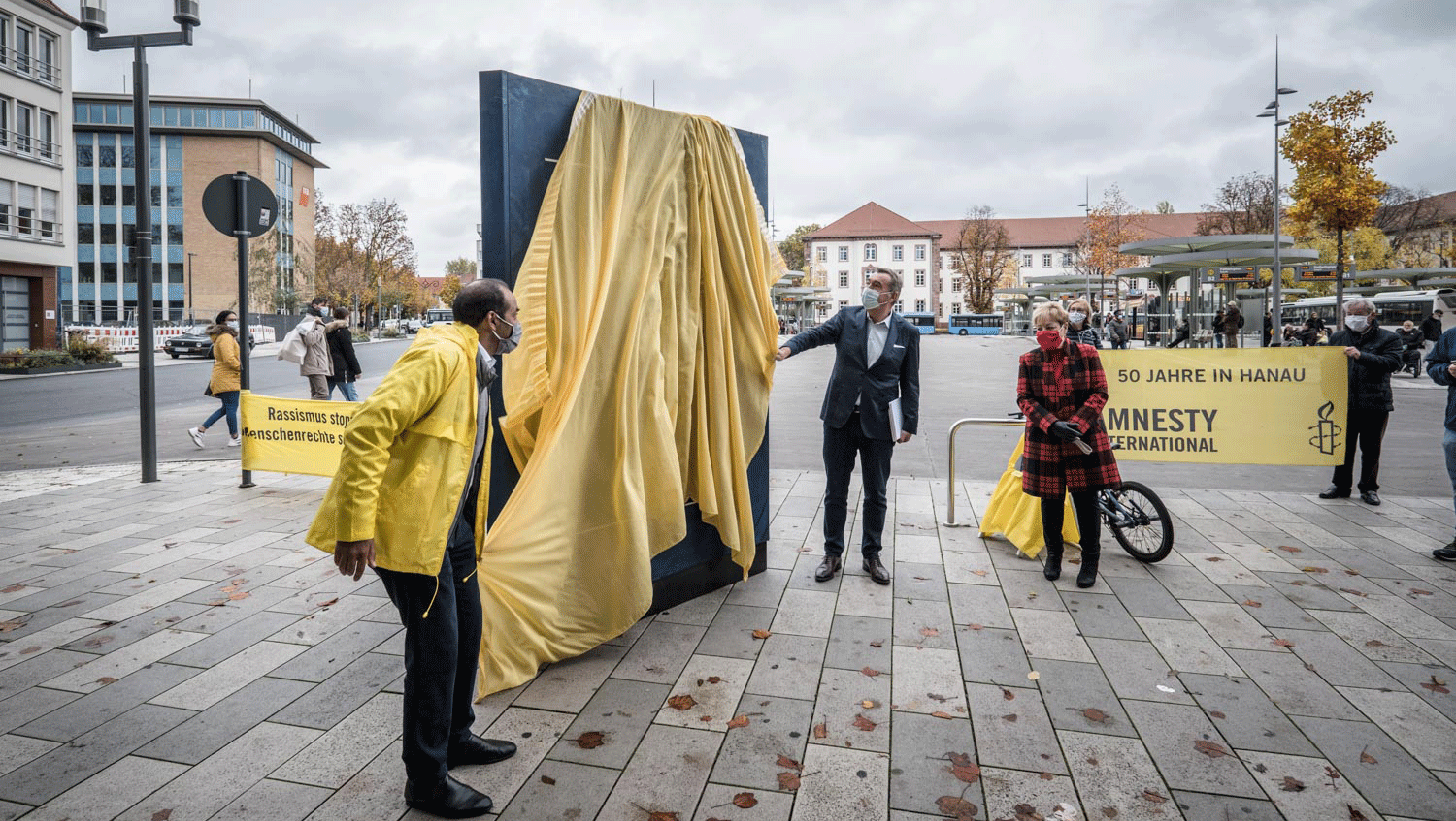 Eine große Version der Menschenrechtserklärung steht auf einem öffentliche Platz, links und rechts davon zwei Personen, sie haben ein gelbes Tuch in der Hand,