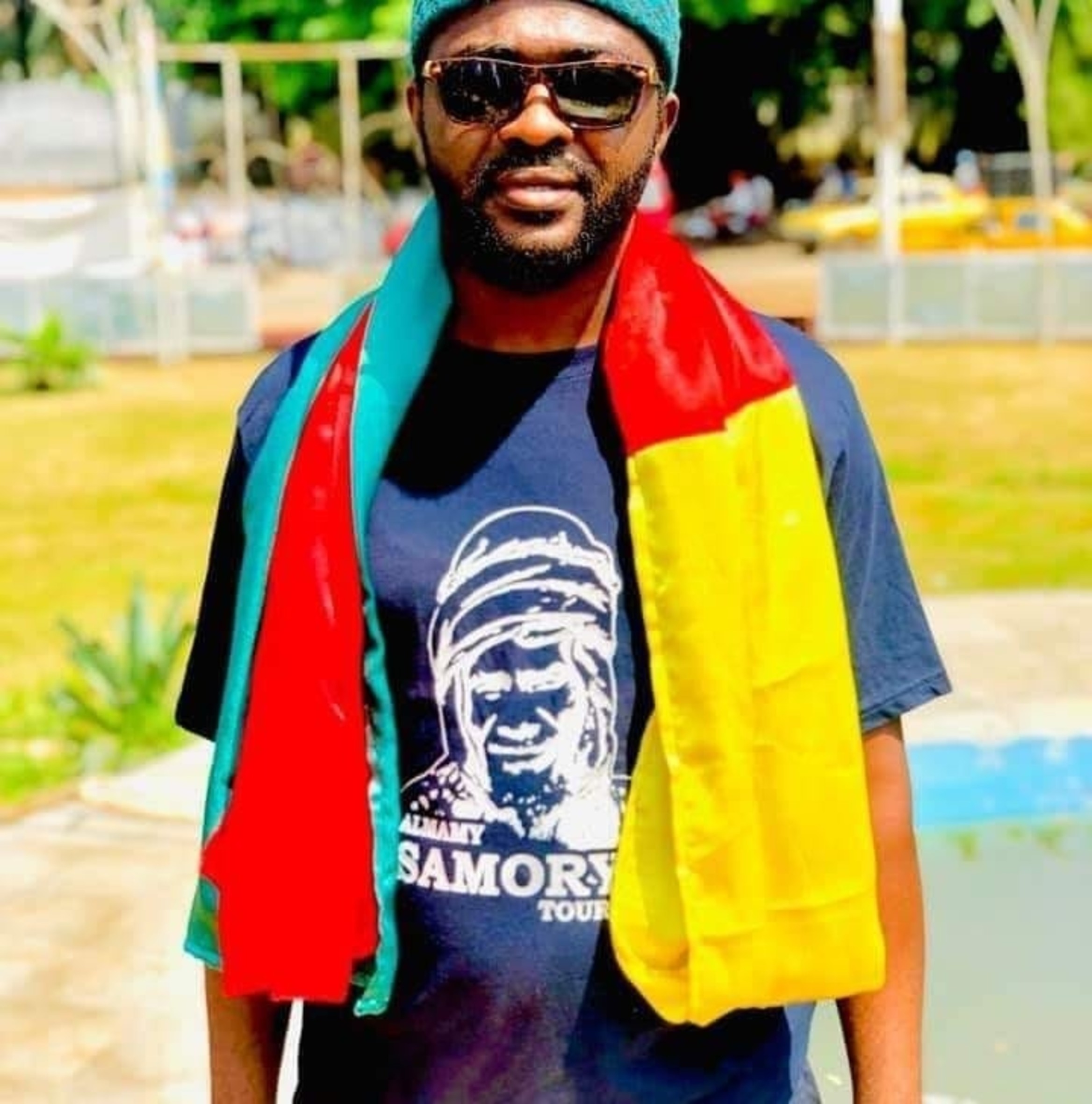 Ein Mann mit Sonnenbrille und grüner Kappe schaut in Richtung Kamera. Er trägt ein dunkelblaues T-Shirt mit einer Skizze von Almamy Samory Touré und diesem Namen in weissen Lettern unter dem Abbild. Um seine Schultern ist ein bunter Schal gelegt.