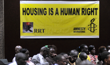 Menschen sitzen am Boden vor einem gelben Plakat, auf dem in schwarzen Lettern "Housing is a human right" steht.
