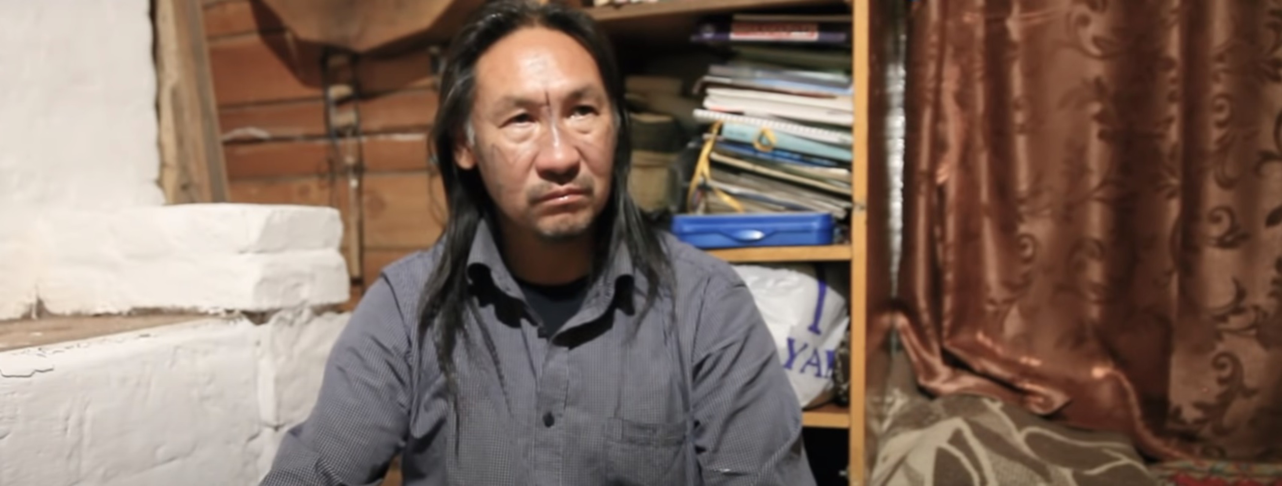 Ein Mann mit schulterlangen schwarzen Haaren und dunkelgrauem Hemd sitzt in einem Zimmer mit Holzwänden. Im Hintergrund hat es einen Aktenstapel.