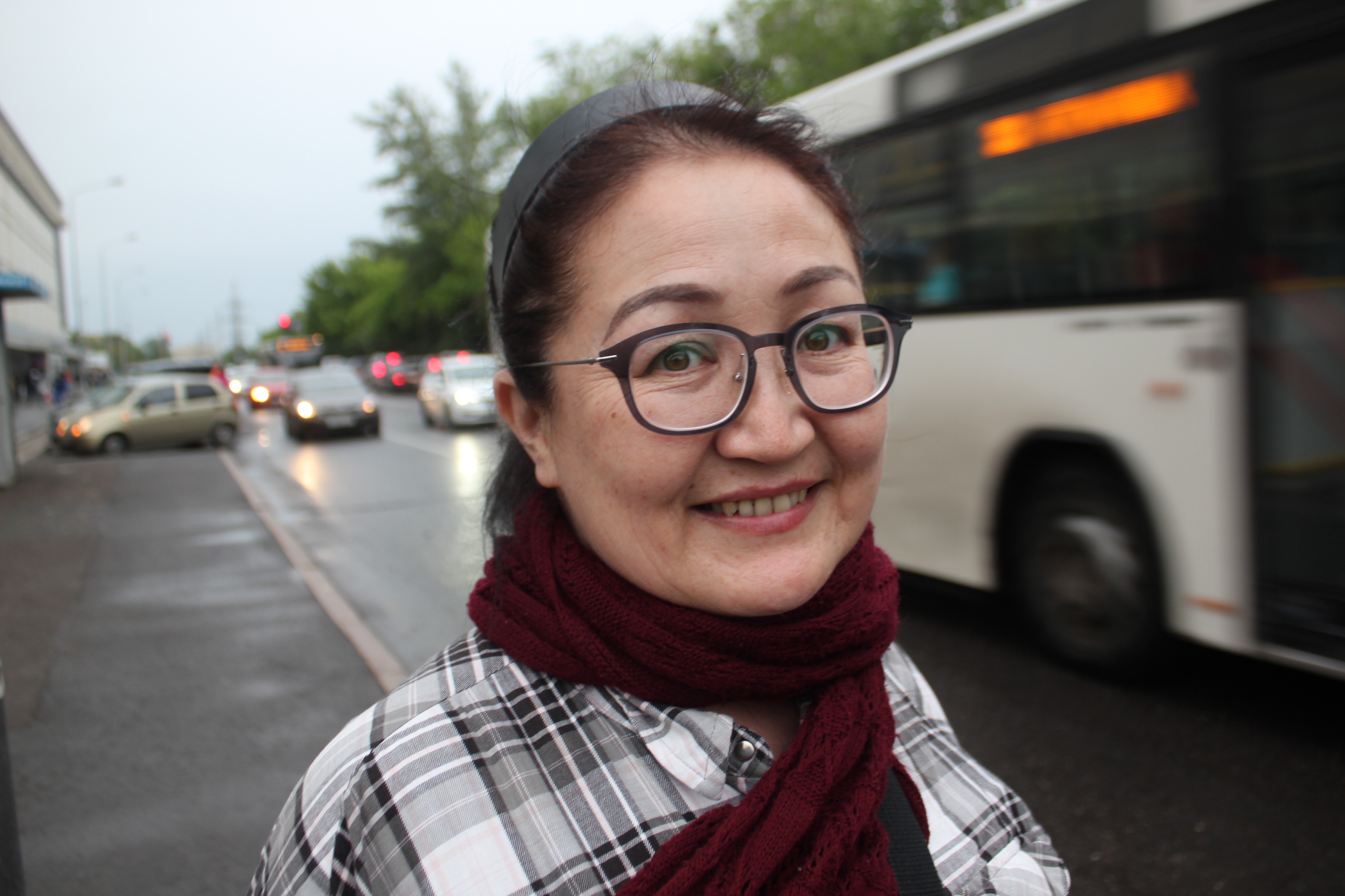 Portrait einer Frau, die in die Kamera lächelt. Sie ist auf der Straße, im Hintergrund ist ein Bus zu sehen.